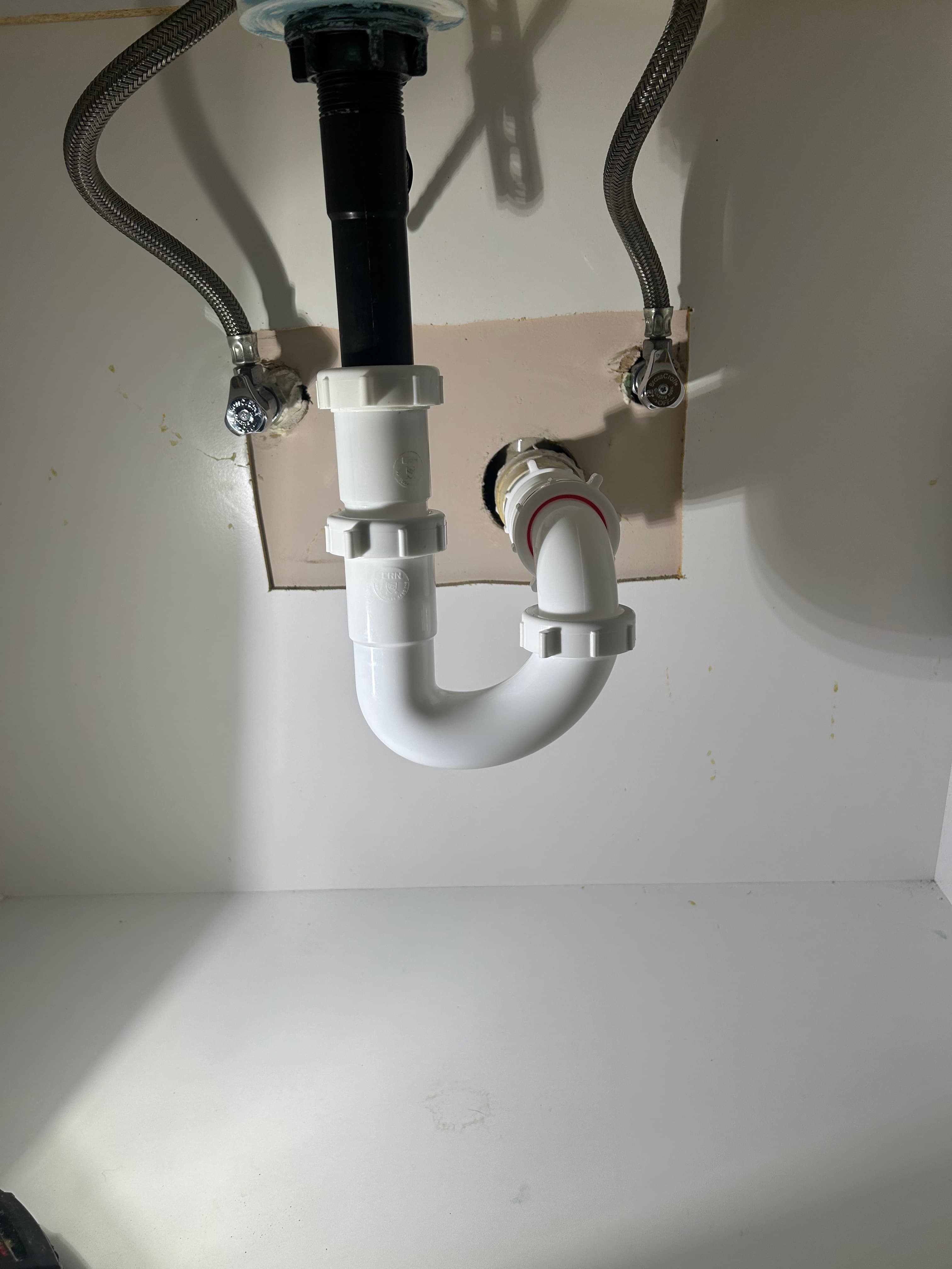 Under-sink plumbing with white plastic P-trap, black tailpiece, and two braided metal water supply lines.