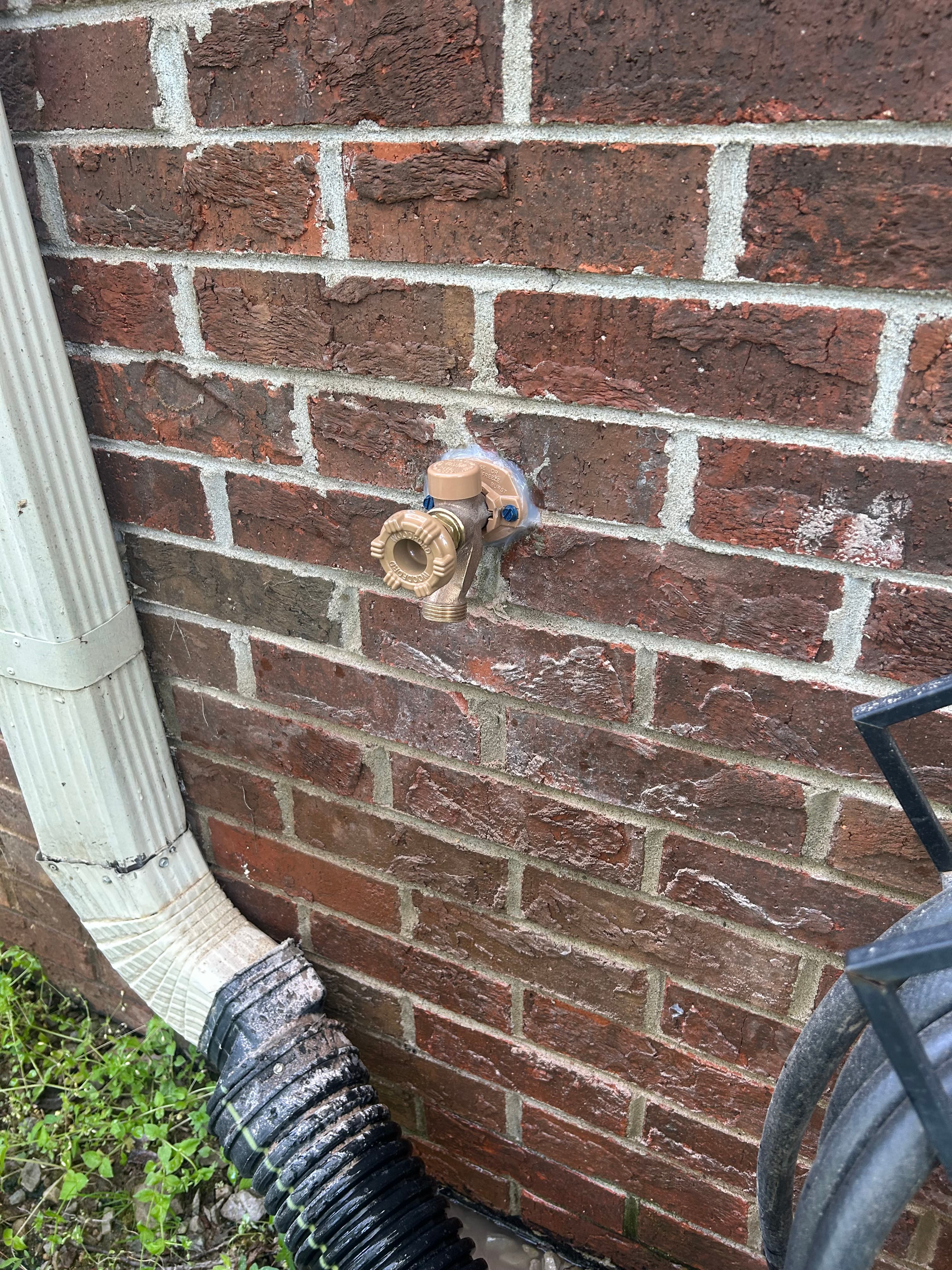 Tan outdoor spigot mounted on a red brick wall next to a white gutter downspout.