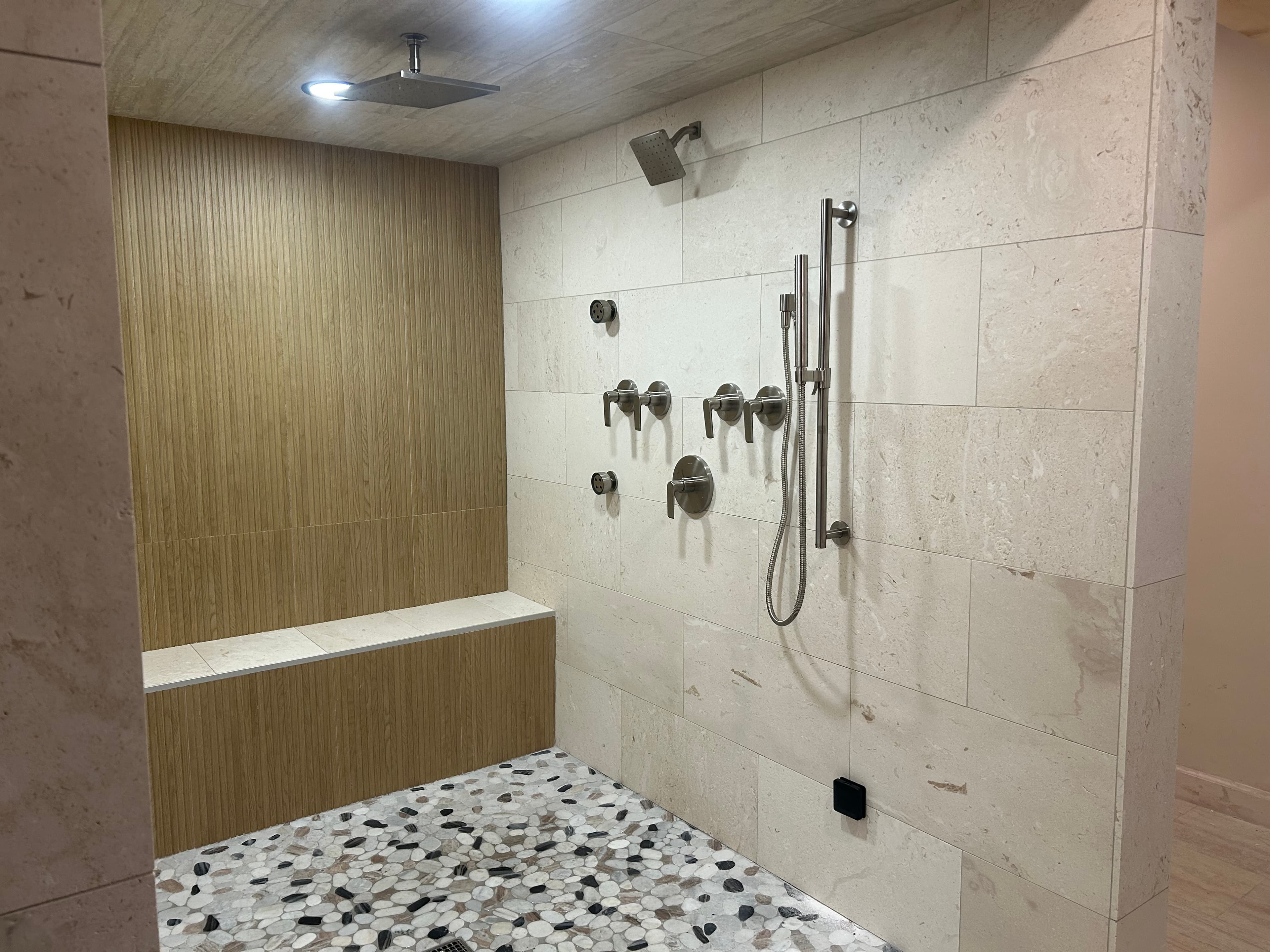 Modern walk-in shower with wood-slat wall, stone tiles, pebble floor, and multiple brushed nickel fixtures.