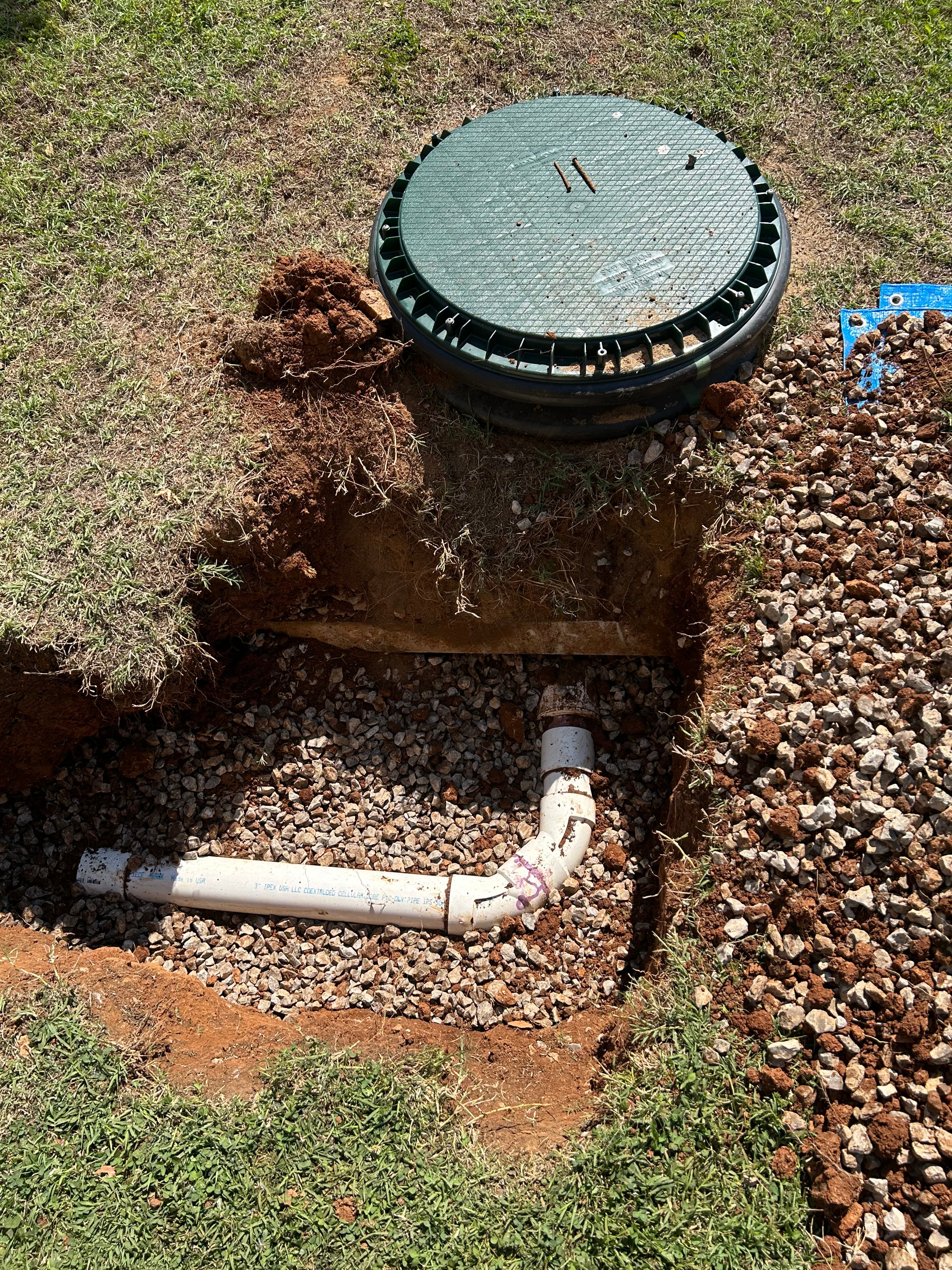 White PVC pipe in a gravel-filled trench beside a green septic tank access cover.