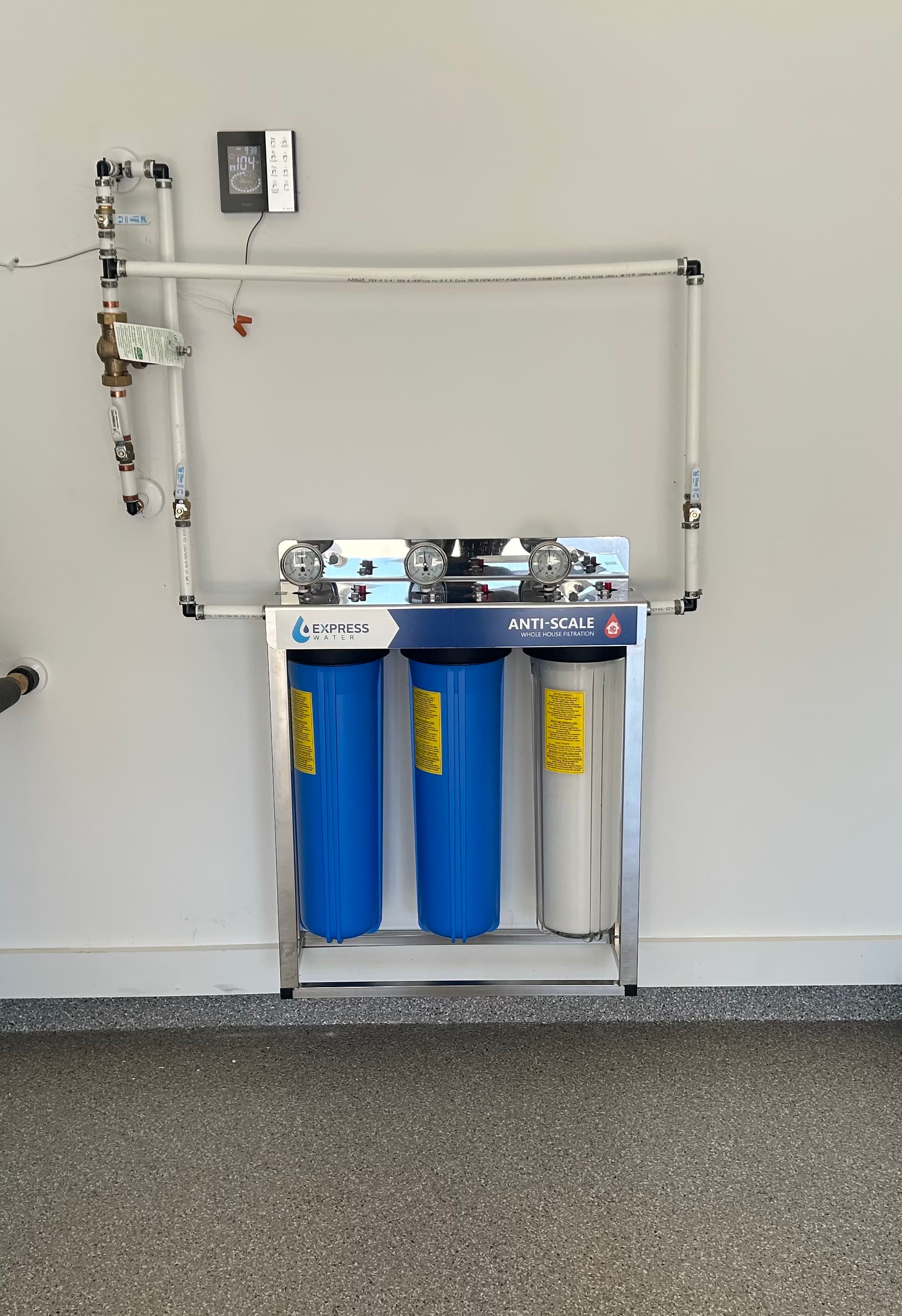 Wall-mounted Express Water three-stage filtration system featuring blue canisters, white piping, and three pressure gauges.